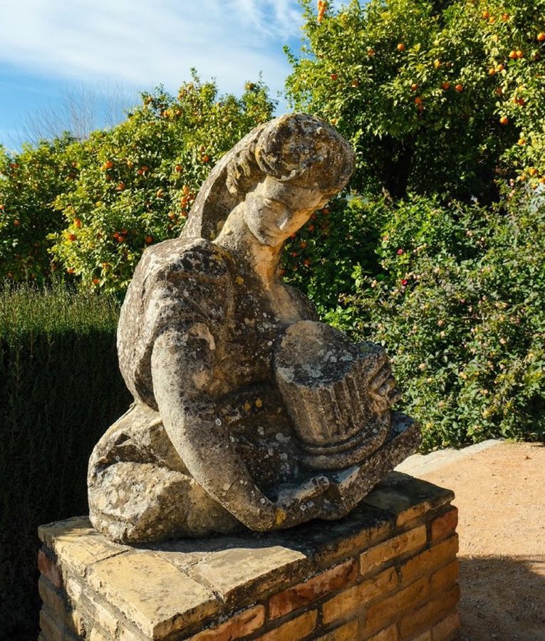 A classical stone sculpture of a seated woman, weathered by time, set against Mediterranean orange trees and blue sky