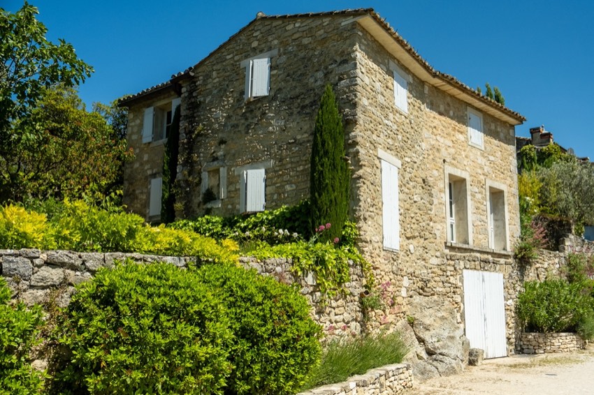 Stone farmhouse facade with white shutters rising above terraced Mediterranean gardens